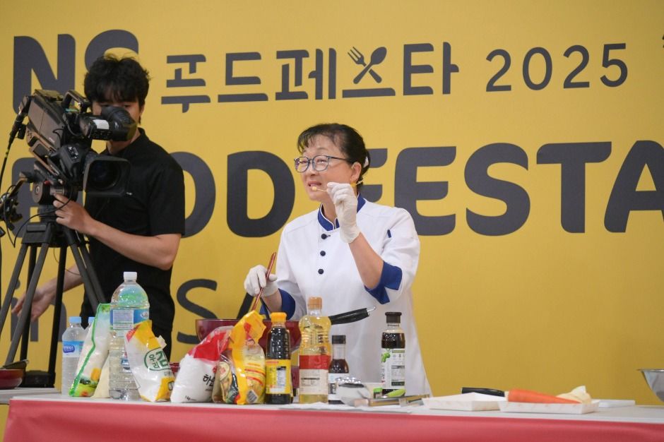 2025년 NS푸드페스타 쿠킹쇼에서 셰프가 재료를 설명하며 조리 시연을 하고 있어요.